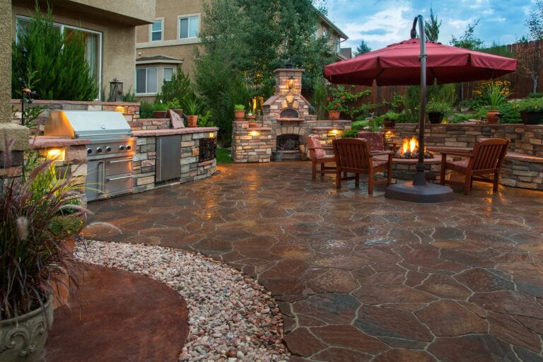 Backyard patio remodel featuring a built-in stone kitchen, wood-fired oven, fire pit, and cozy seating area under a red umbrella.